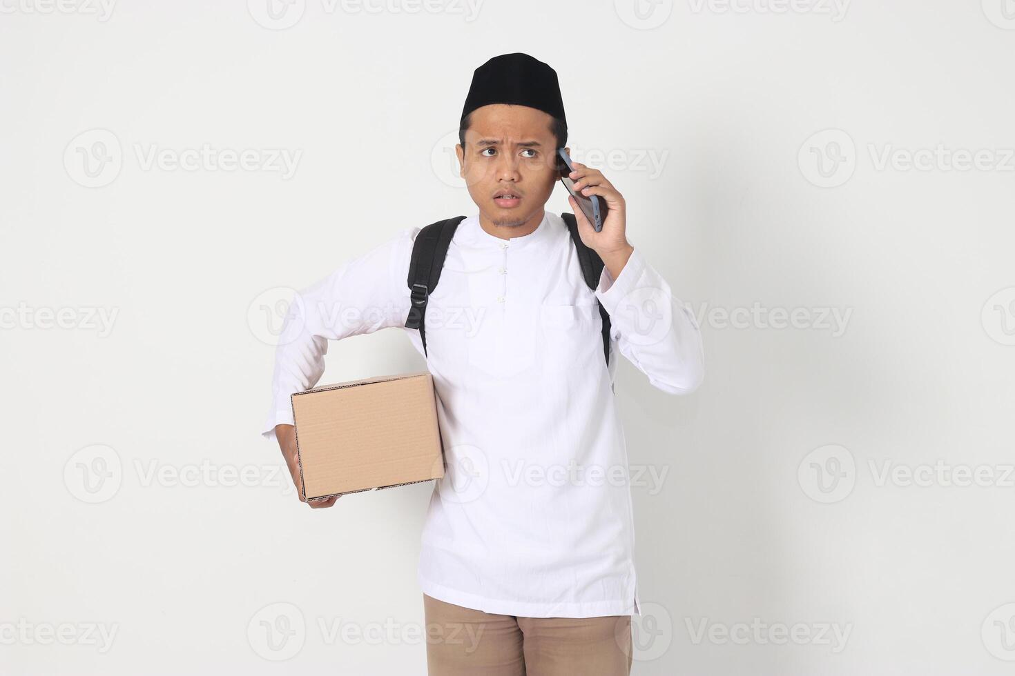 Portrait of worried Asian muslim carrying cardboard box while having having call on mobile phone, showing sad expression. Going home for Eid Mubarak. Isolated image on white background photo