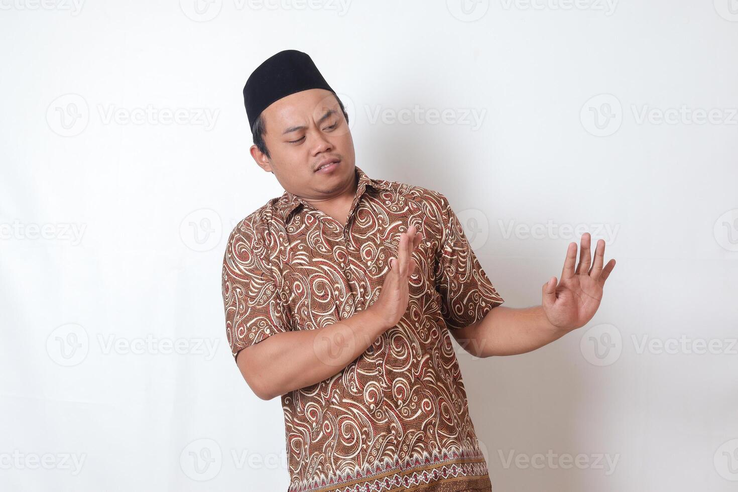 Portrait of unpleasant Asian man wearing batik shirt and songkok forming a hand gesture to avoid something. Advertising concept. Isolated image on gray background photo