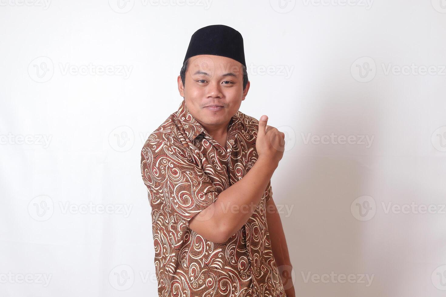 Portrait of excited Asian man wearing batik shirt and songkok smiling and looking at camera, making thumbs up hand gesture. Isolated image on gray background photo