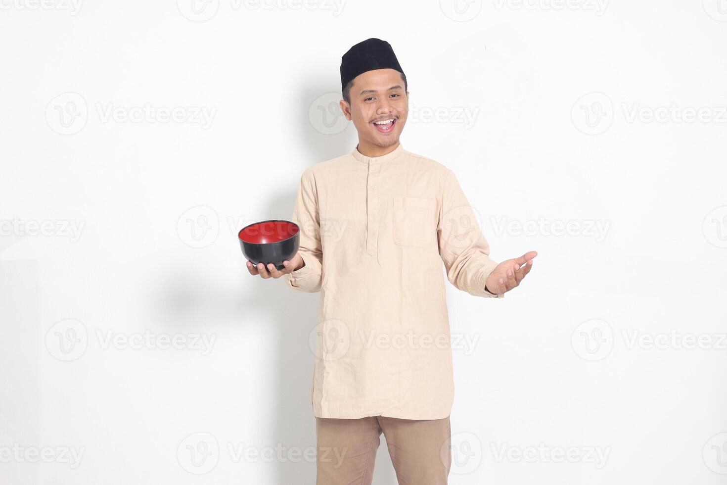 Portrait of excited Asian muslim man in koko shirt with skullcap showing and holding an empty plate. Bowl template for food brand. Isolated on white background photo