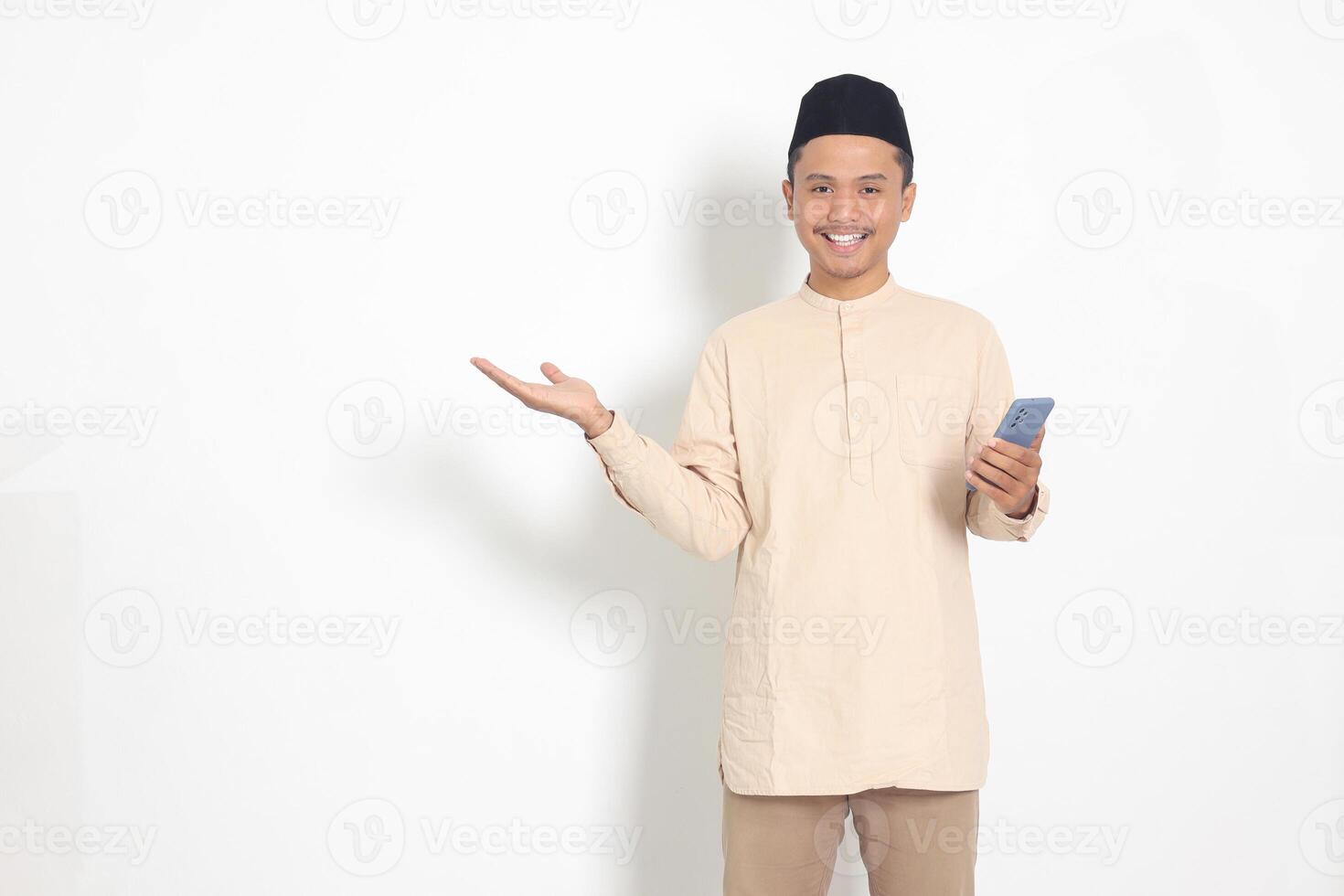 Portrait of attractive Asian muslim man in koko shirt with skullcap holding mobile phone, pointing and showing product with finger. Social media concept. Isolated image on white background photo