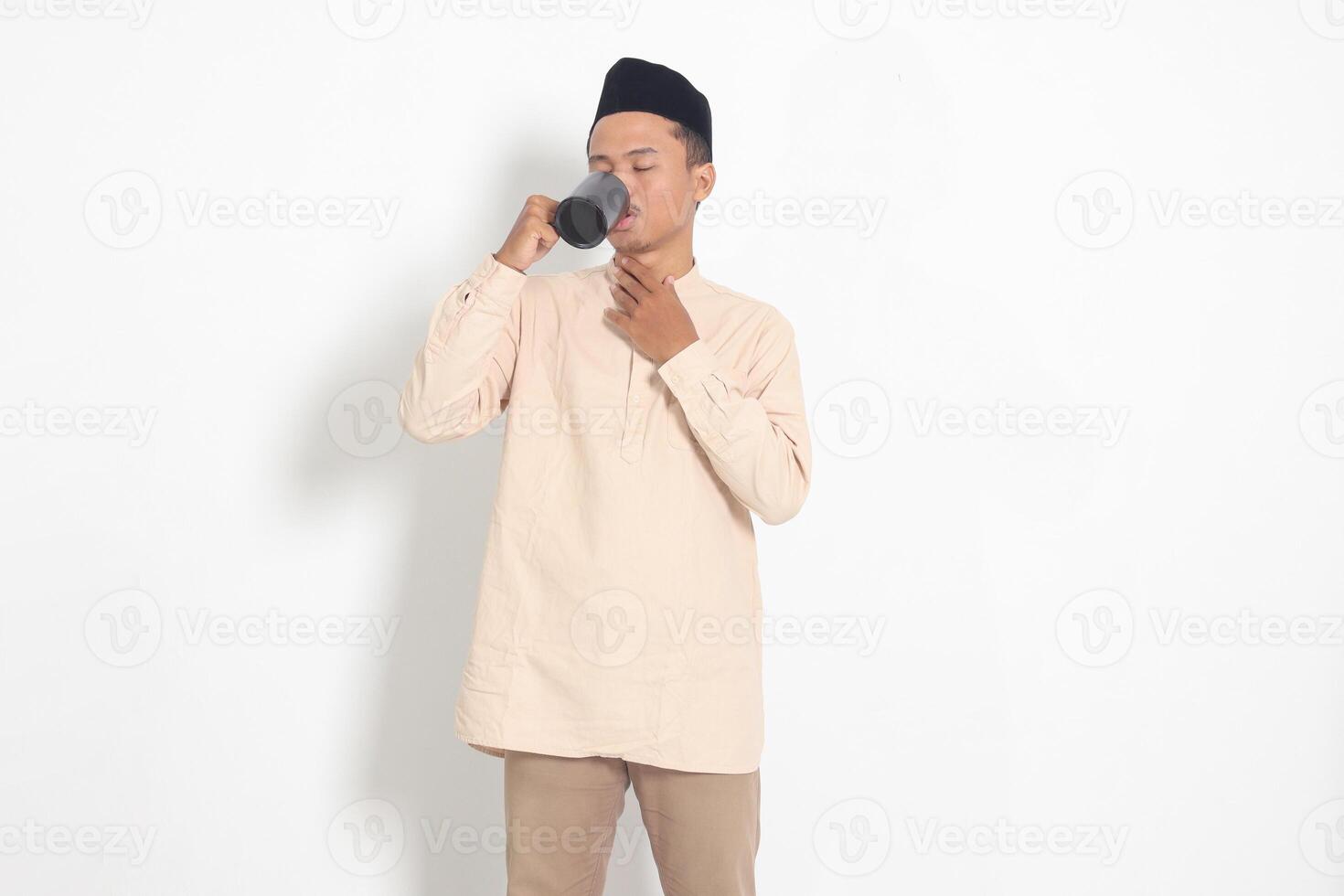 Portrait of attractive Asian muslim man in koko shirt with peci holding a mug, drinking water, quenching thirst after fasting. Isolated image on white background photo