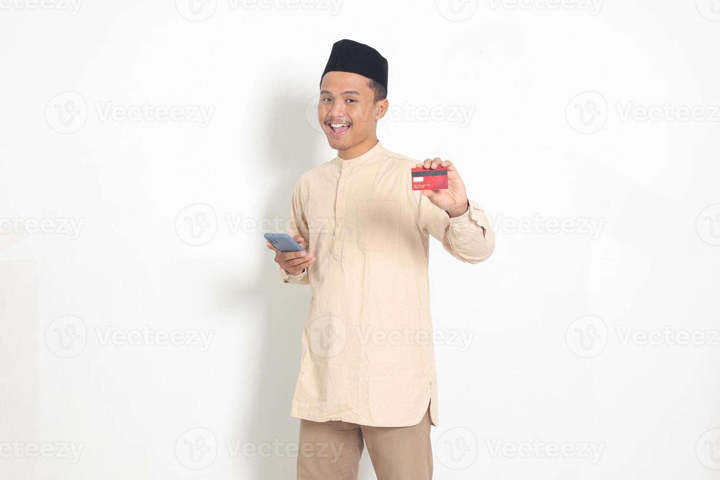 Portrait of attractive Asian muslim man in koko shirt with skullcap holding a mobile phone and presenting credit card. Isolated image on white background photo