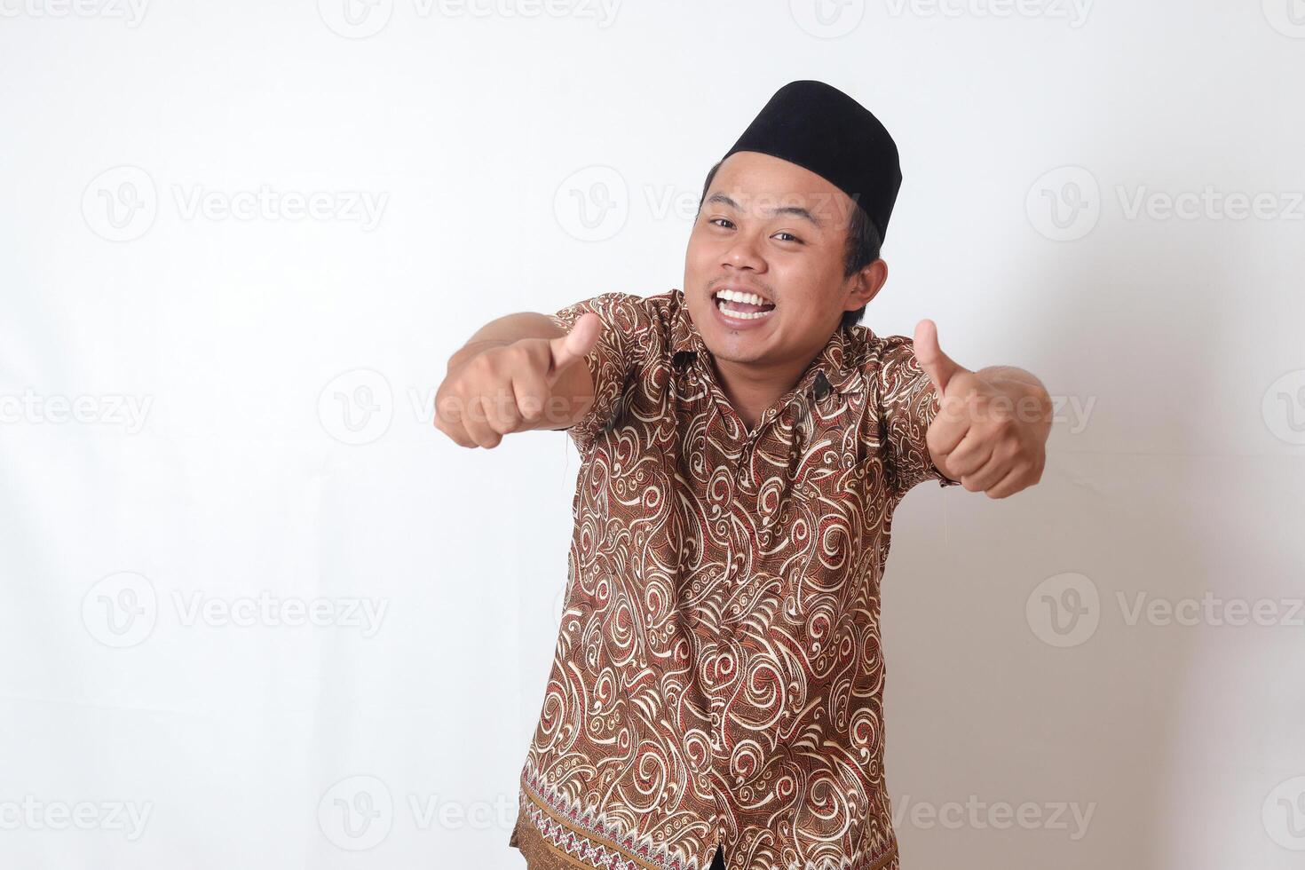 retrato de emocionado asiático hombre vistiendo batik camisa y Songkok sonriente y mirando a cámara, haciendo pulgares arriba mano gesto. aislado imagen en gris antecedentes foto