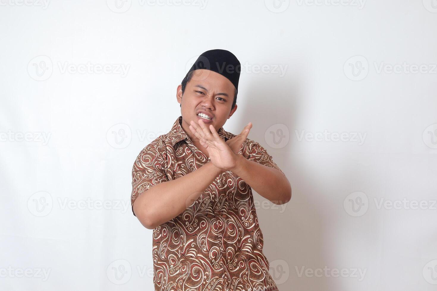 Portrait of excited Asian man wearing batik shirt and songkok  showing X sign of hands, refusing an invitation from someone, making prohibitive gesture. Isolated image on gray background photo