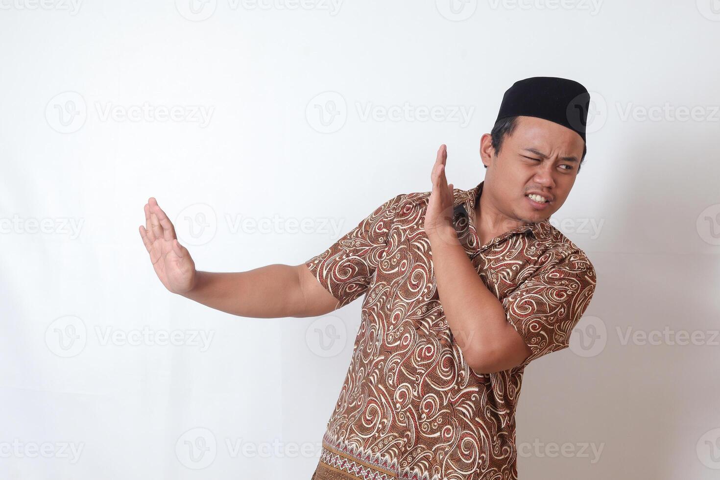Portrait of unpleasant Asian man wearing batik shirt and songkok forming a hand gesture to avoid something. Advertising concept. Isolated image on gray background photo