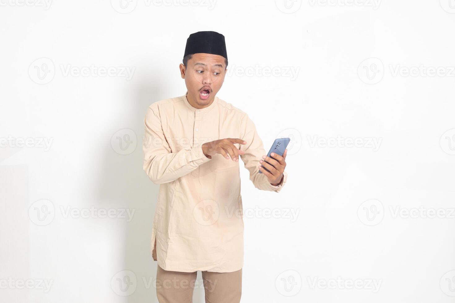 Portrait of surprised Asian muslim man in koko shirt with skullcap holding mobile phone, showing shocked face expression. Advertising and social media concept. Isolated image on white background photo