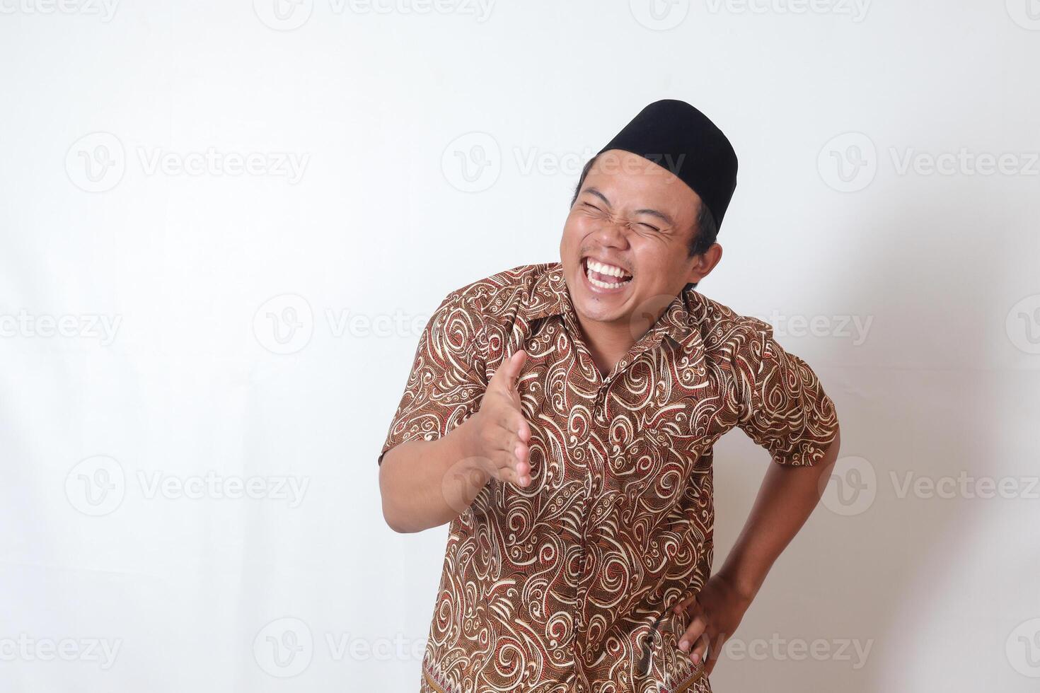 retrato de emocionado asiático hombre vistiendo batik camisa y Songkok alcanzando fuera su mano para un mano agitar, acogedor alguien. aislado imagen en gris antecedentes foto
