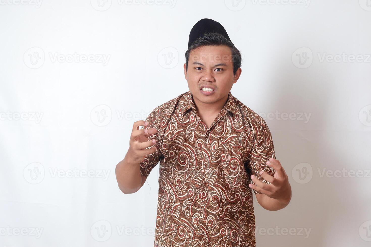 Portrait of annoyed Asian man wearing batik shirt and songkok making angry hand gesture with fingers. Isolated image on gray background photo