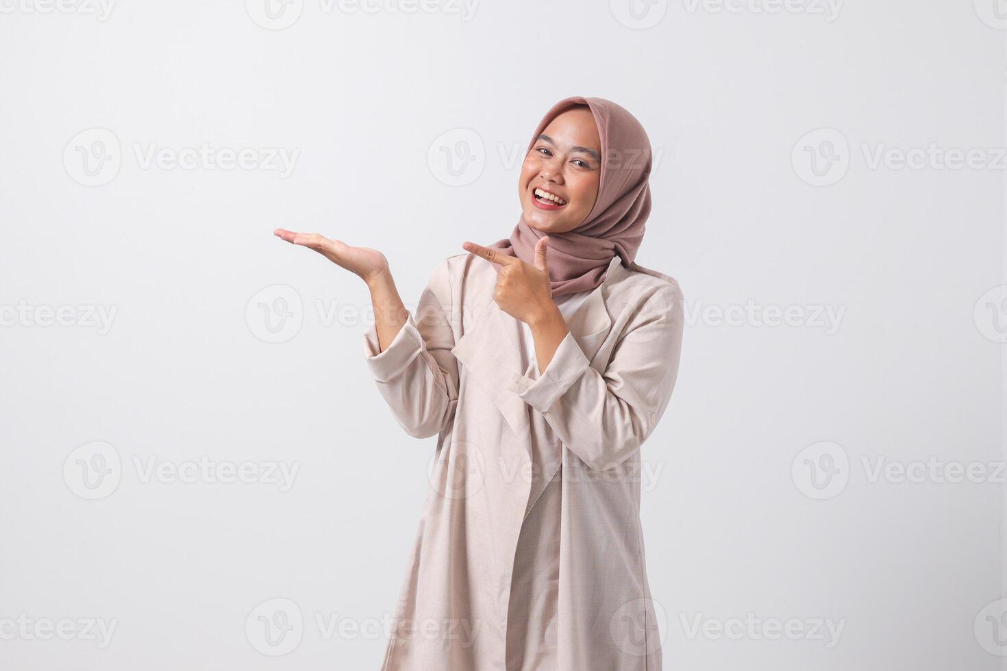 Portrait of excited Asian hijab woman in casual suit pointing and showing product in her side with finger. Businesswoman concept. Isolated image on white background photo