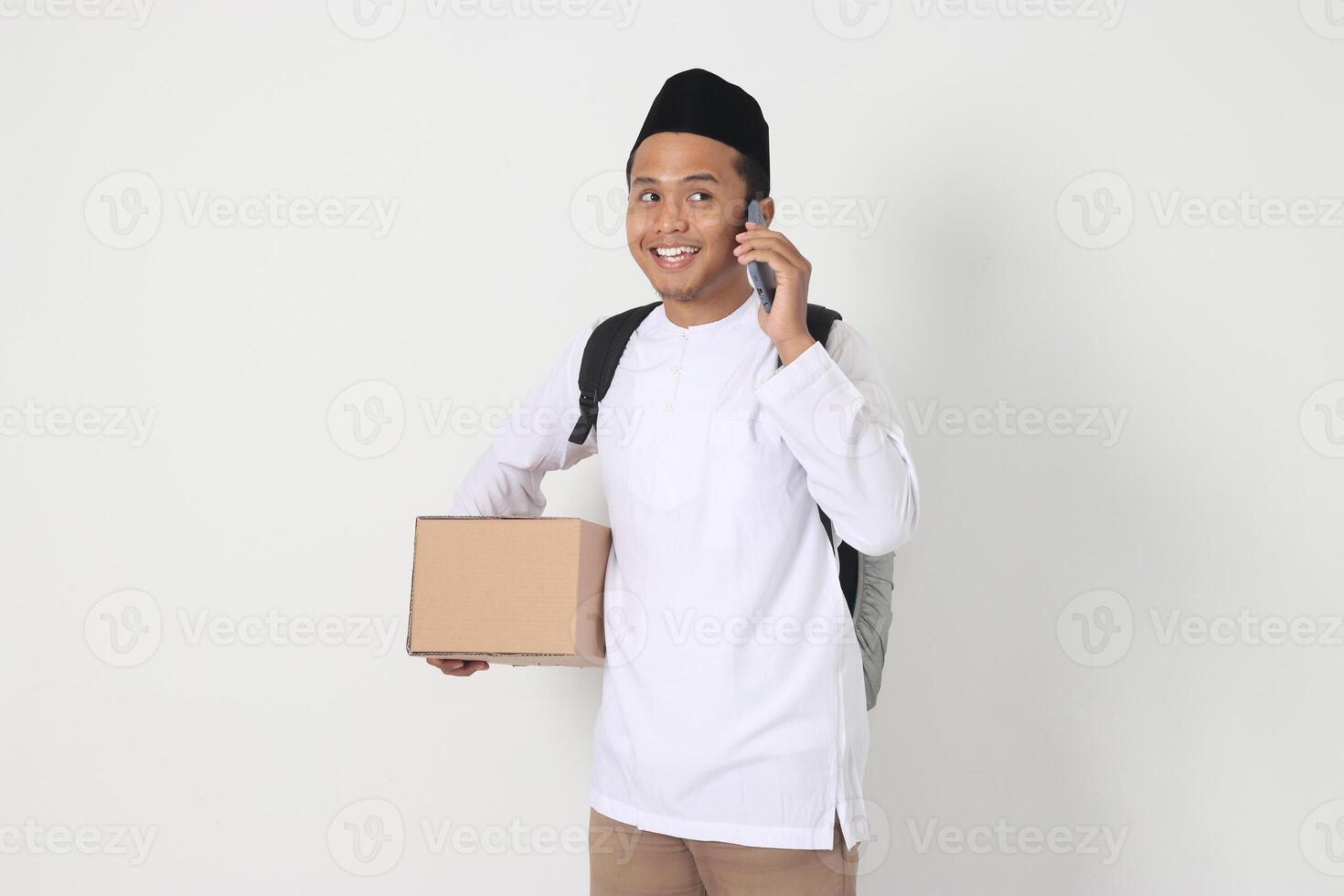 Portrait of happy Asian muslim man in koko shirt with peci carrying cardboard box while talking to his friend and family on cellphone. Going home for Eid Mubarak. Isolated image on white background photo