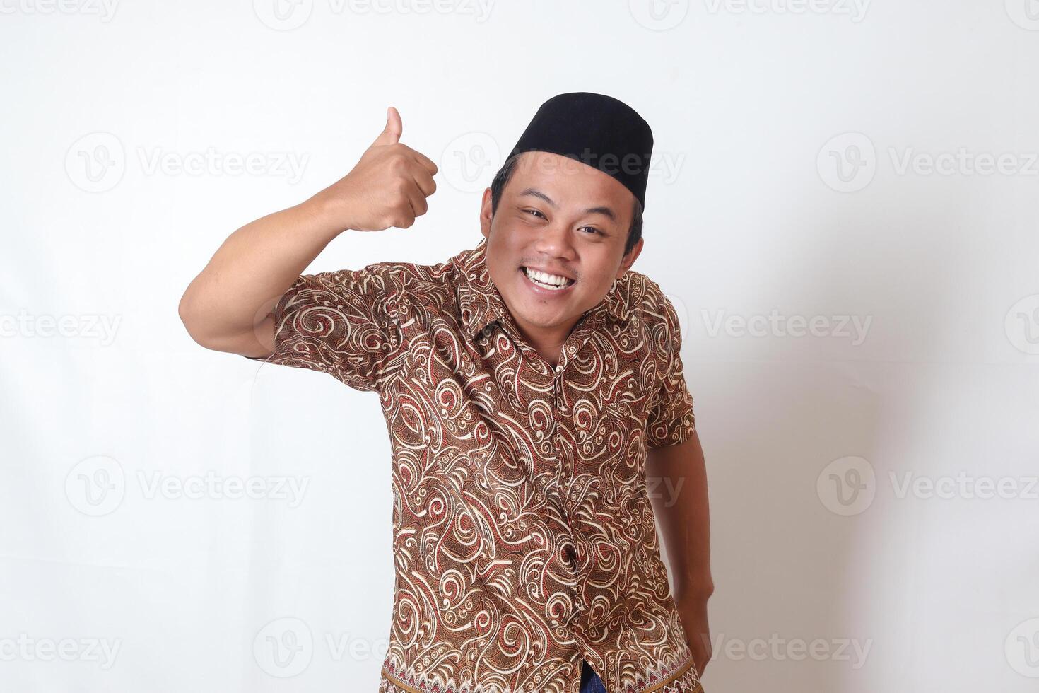 Portrait of excited Asian man wearing batik shirt and songkok smiling and looking at camera, making thumbs up hand gesture. Isolated image on gray background photo