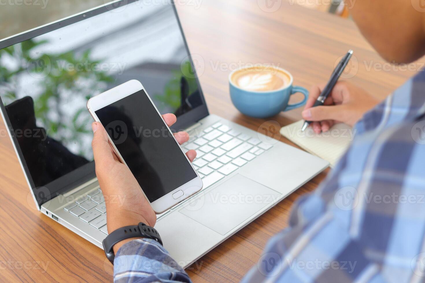 Close up of male hand writing on note book with pen, showing blank screen of laptop and smartphone. Working in cafe concept with a cup of coffee photo