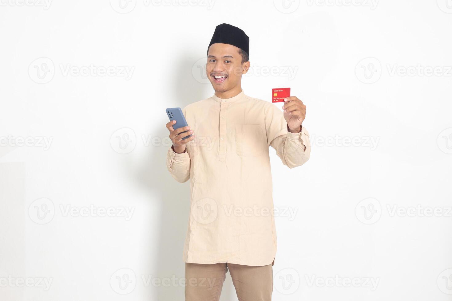 Portrait of attractive Asian muslim man in koko shirt with skullcap holding a mobile phone and presenting credit card. Isolated image on white background photo