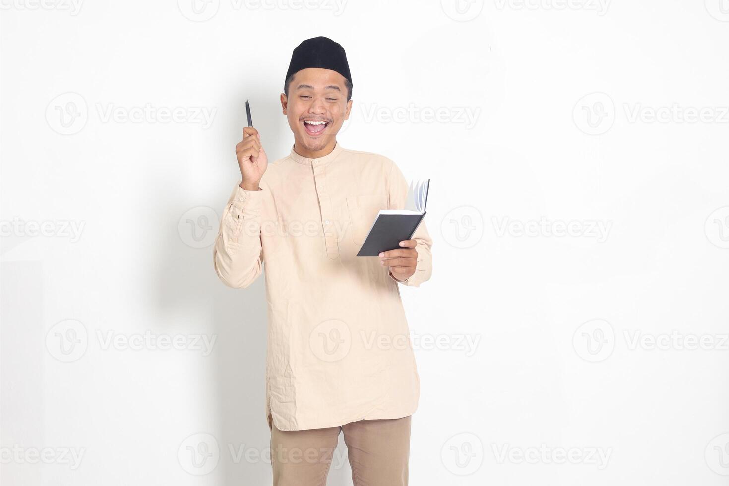 Portrait of attractive Asian muslim man in koko shirt with peci reading a book, telling that he has an idea while pointing finger and pen. Isolated image on white background photo