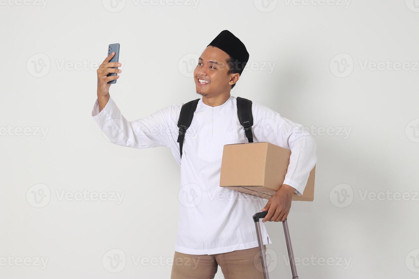 Portrait of excited Asian muslim man carrying cardboard box and suitcase while taking picture of himself or seflie and video calling. Going home for Eid Mubarak. Isolated image on white background photo