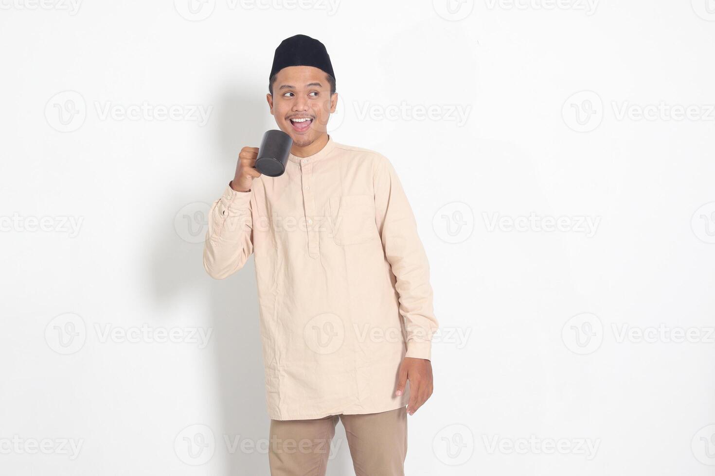 Portrait of attractive Asian muslim man in koko shirt with peci holding a mug, drinking water, quenching thirst after fasting. Isolated image on white background photo
