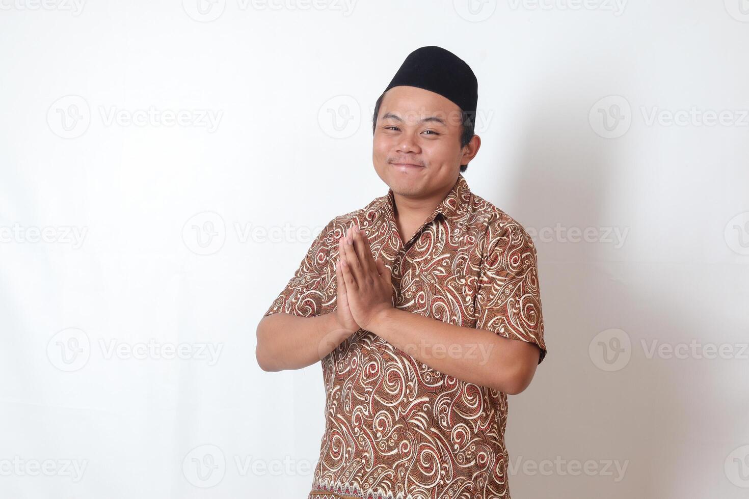 Portrait of smiling Asian man wearing batik shirt and songkok showing apologize and welcome hand gesture. Ramadan advertising concept. Isolated image on gray background photo