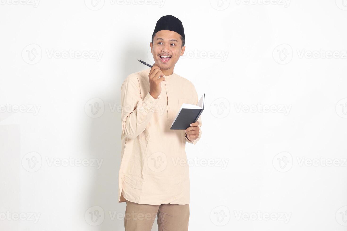 Portrait of excited muslim man in koko shirt with peci reading a notebook. Happy Asian guy pointing to the side with finger. Isolated image on white background photo