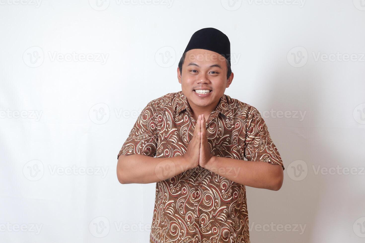 Portrait of smiling Asian man wearing batik shirt and songkok showing apologize and welcome hand gesture. Ramadan advertising concept. Isolated image on gray background photo