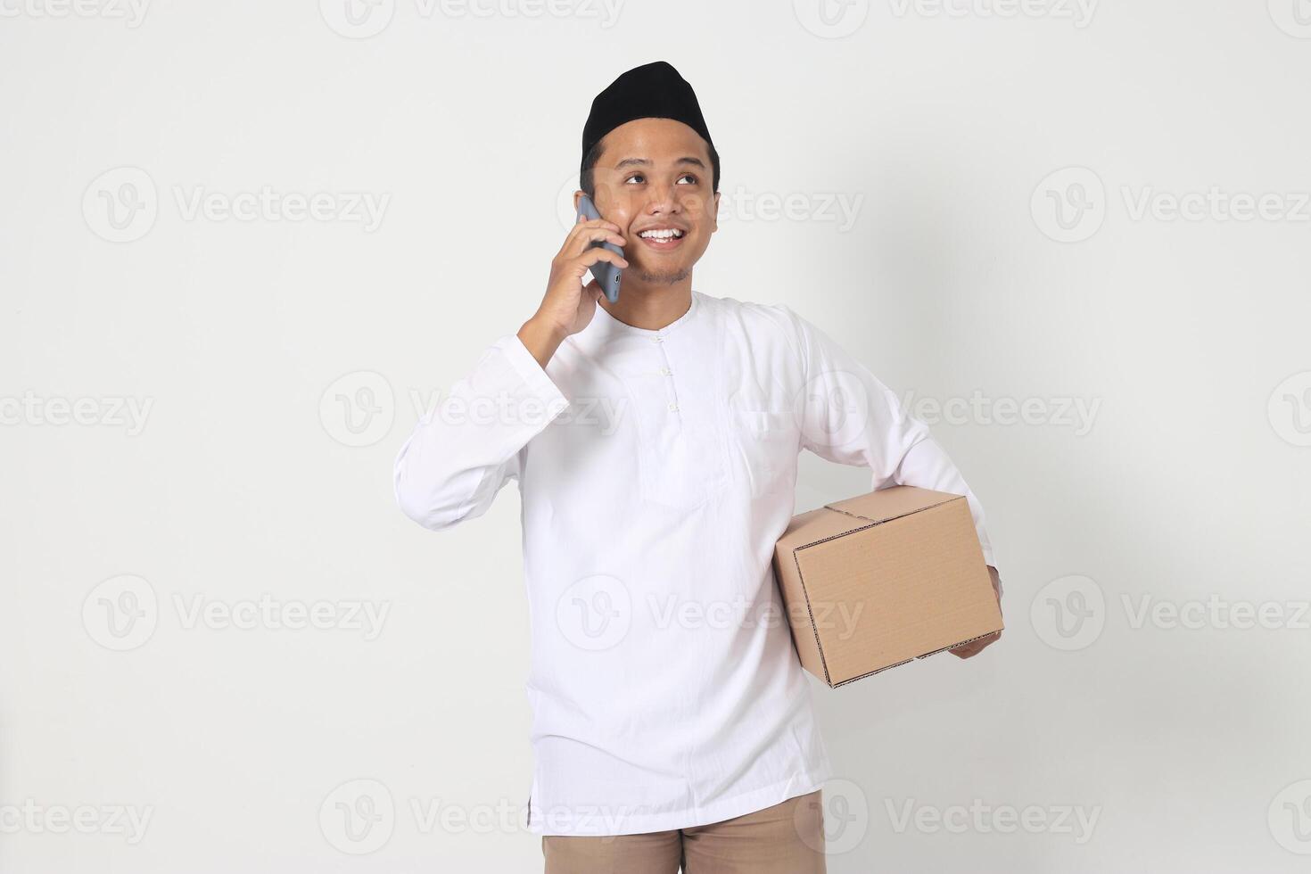 Portrait of happy Asian muslim man in koko shirt with peci carrying cardboard box while having having conversation on mobile phone. Going home for Eid Mubarak. Isolated image on white background photo