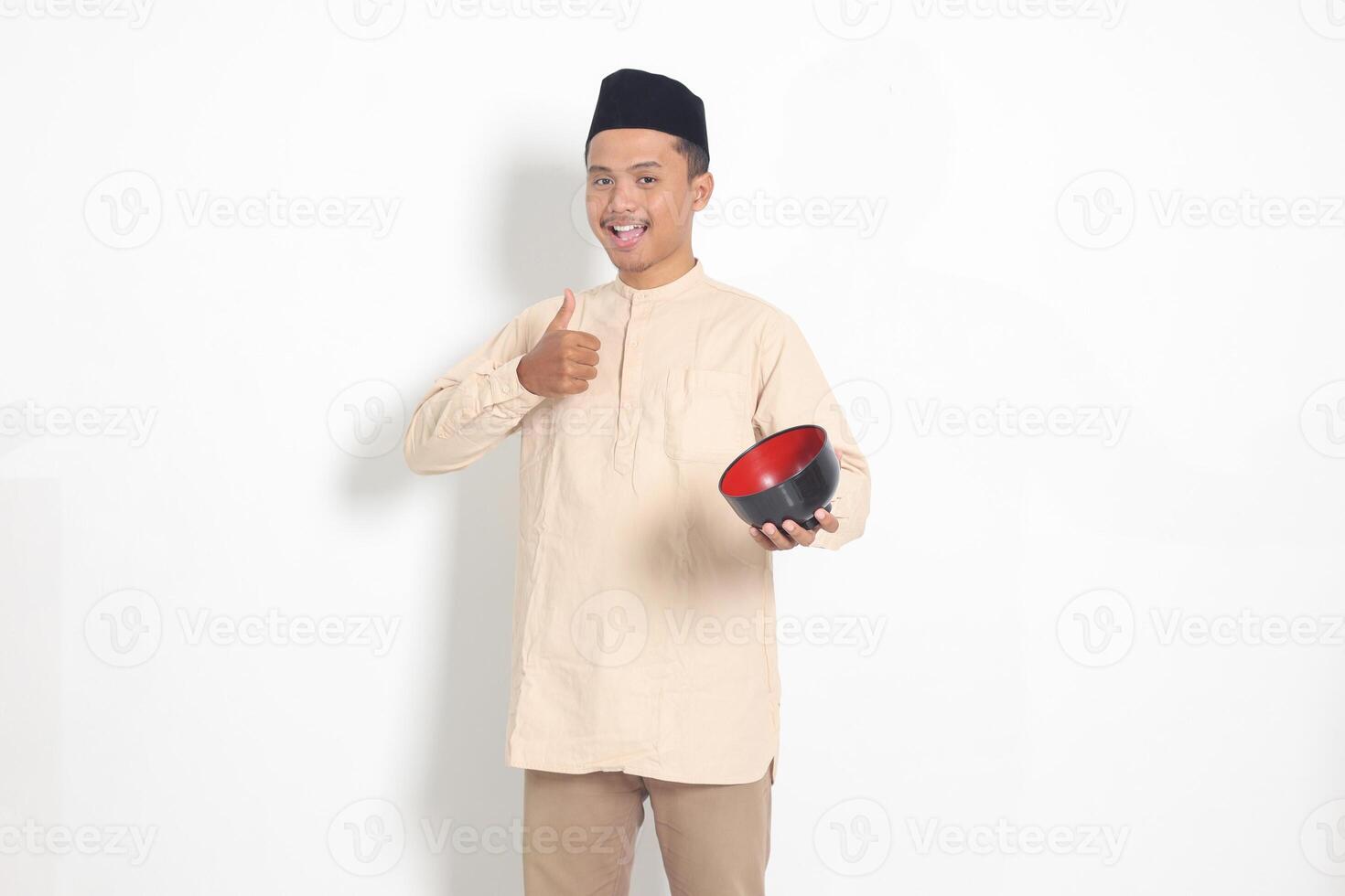 Portrait of excited Asian muslim man in koko shirt with skullcap showing and holding an empty plate. Bowl template for food brand. Isolated on white background photo