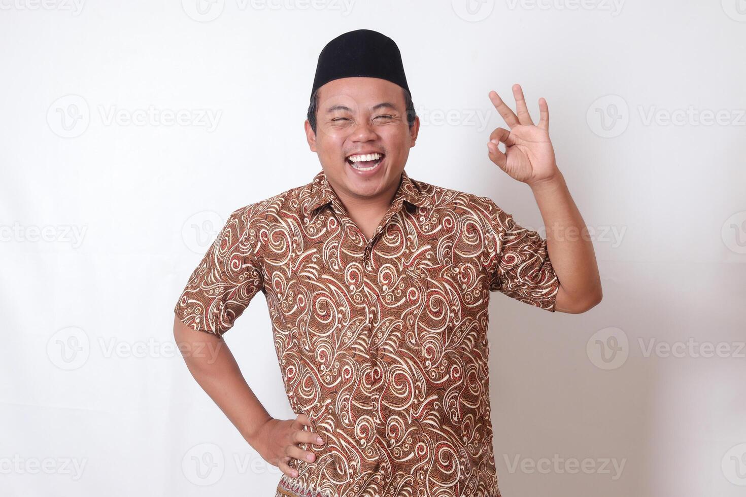 Portrait of excited Asian man wearing batik shirt and songkok showing ok hand gesture and smiling looking at camera. Isolated image on gray background photo