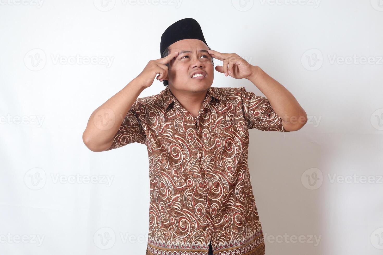 retrato de sufrimiento asiático hombre vistiendo batik camisa y Songkok teniendo dolor de cabeza mientras conmovedor su frente área. aislado imagen en gris antecedentes foto