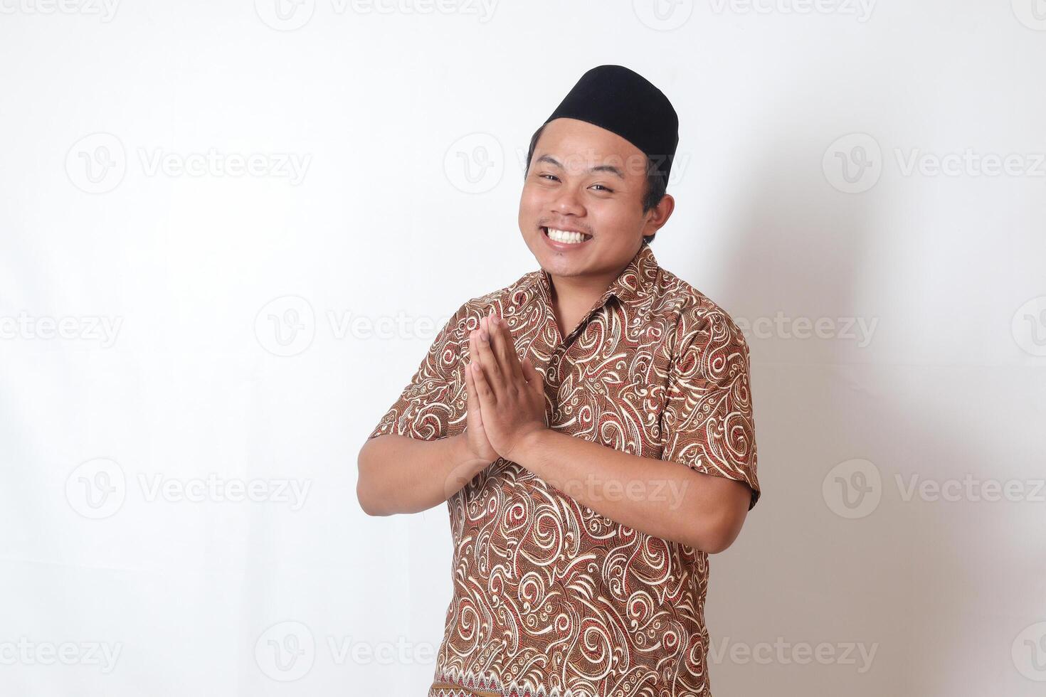 retrato de sonriente asiático hombre vistiendo batik camisa y Songkok demostración pedir disculpas y Bienvenido mano gesto. Ramadán publicidad concepto. aislado imagen en gris antecedentes foto