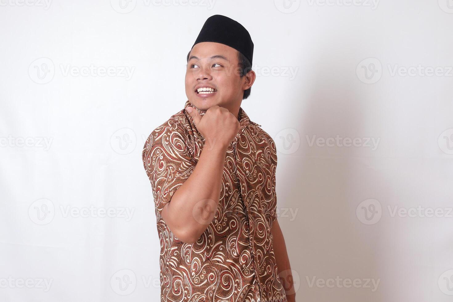Portrait of excited Asian man wearing batik shirt and songkok smiling and looking at the camera pointing with two hands and fingers to the side. Isolated image on gray background photo