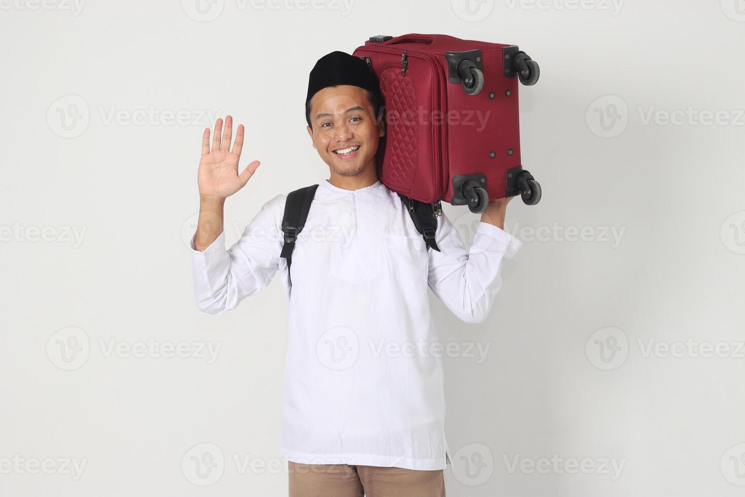 Portrait of attractive Asian muslim man carrying suitcase while saying hi to greet someone. Going home for Eid Mubarak. Isolated image on white background photo