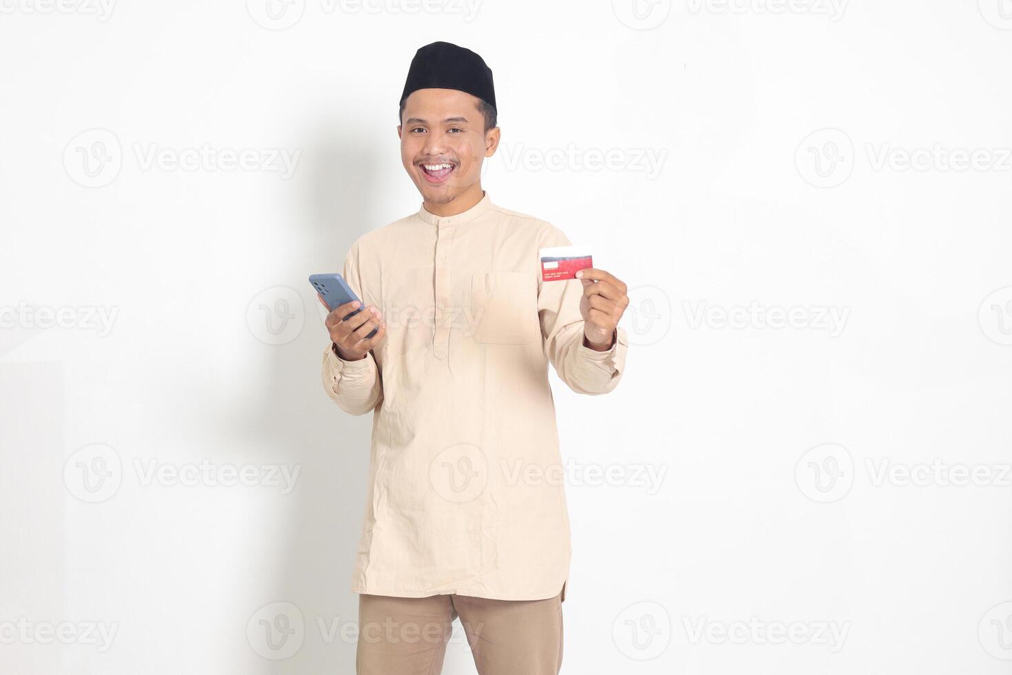 Portrait of attractive Asian muslim man in koko shirt with skullcap holding a mobile phone and presenting credit card. Isolated image on white background photo
