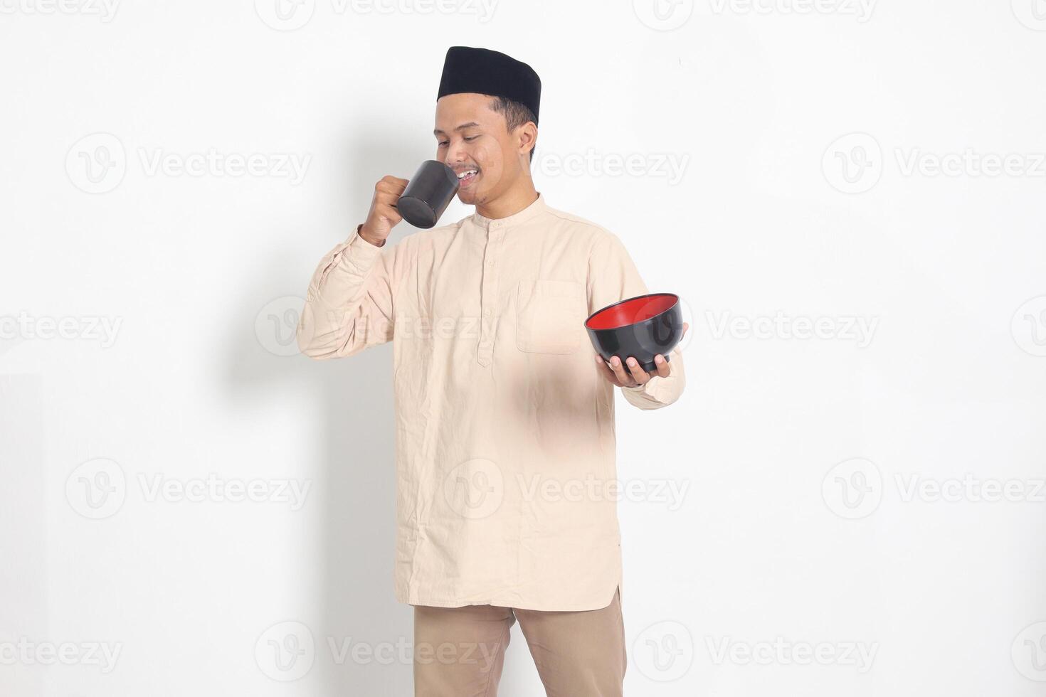 Portrait of excited Asian muslim man in koko shirt with skullcap holding a mug and empty bowl. Bowl template for food brand. Isolated image on white background photo