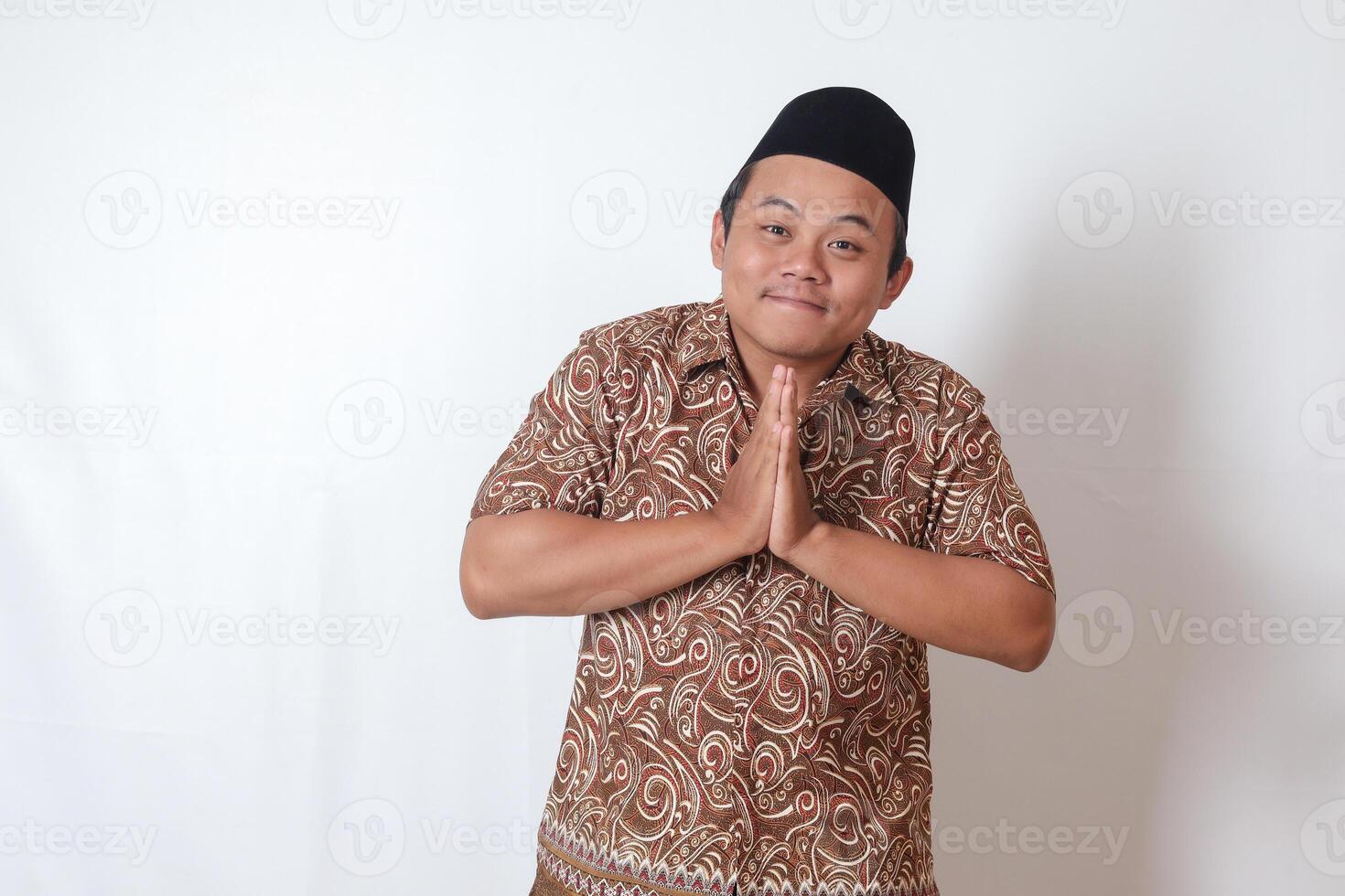 Portrait of smiling Asian man wearing batik shirt and songkok showing apologize and welcome hand gesture. Ramadan advertising concept. Isolated image on gray background photo