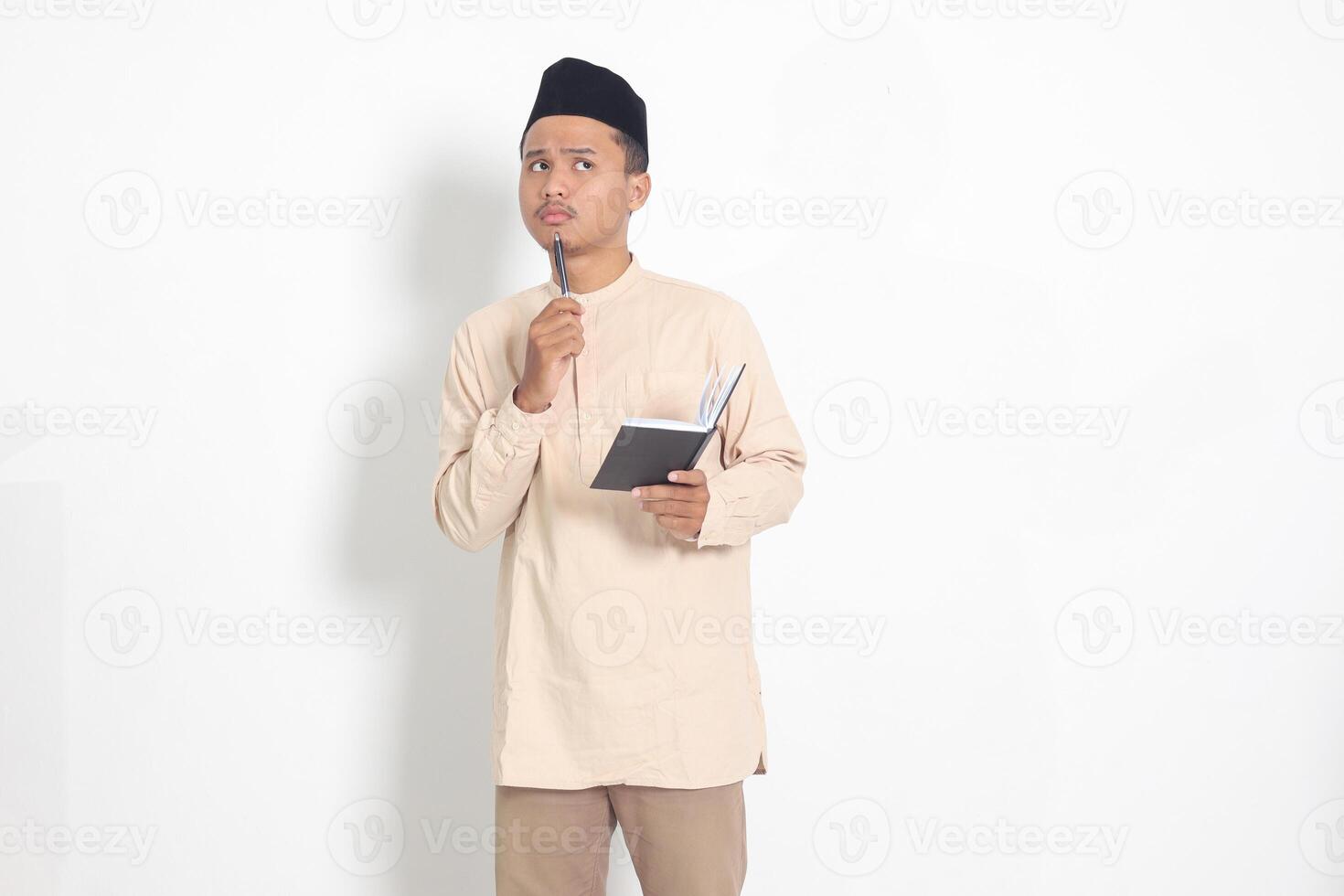 Portrait of confused Asian muslim man in koko shirt with peci reading a book, thinking about an idea with hand holding pen on chin, looking away. Isolated image on white background photo