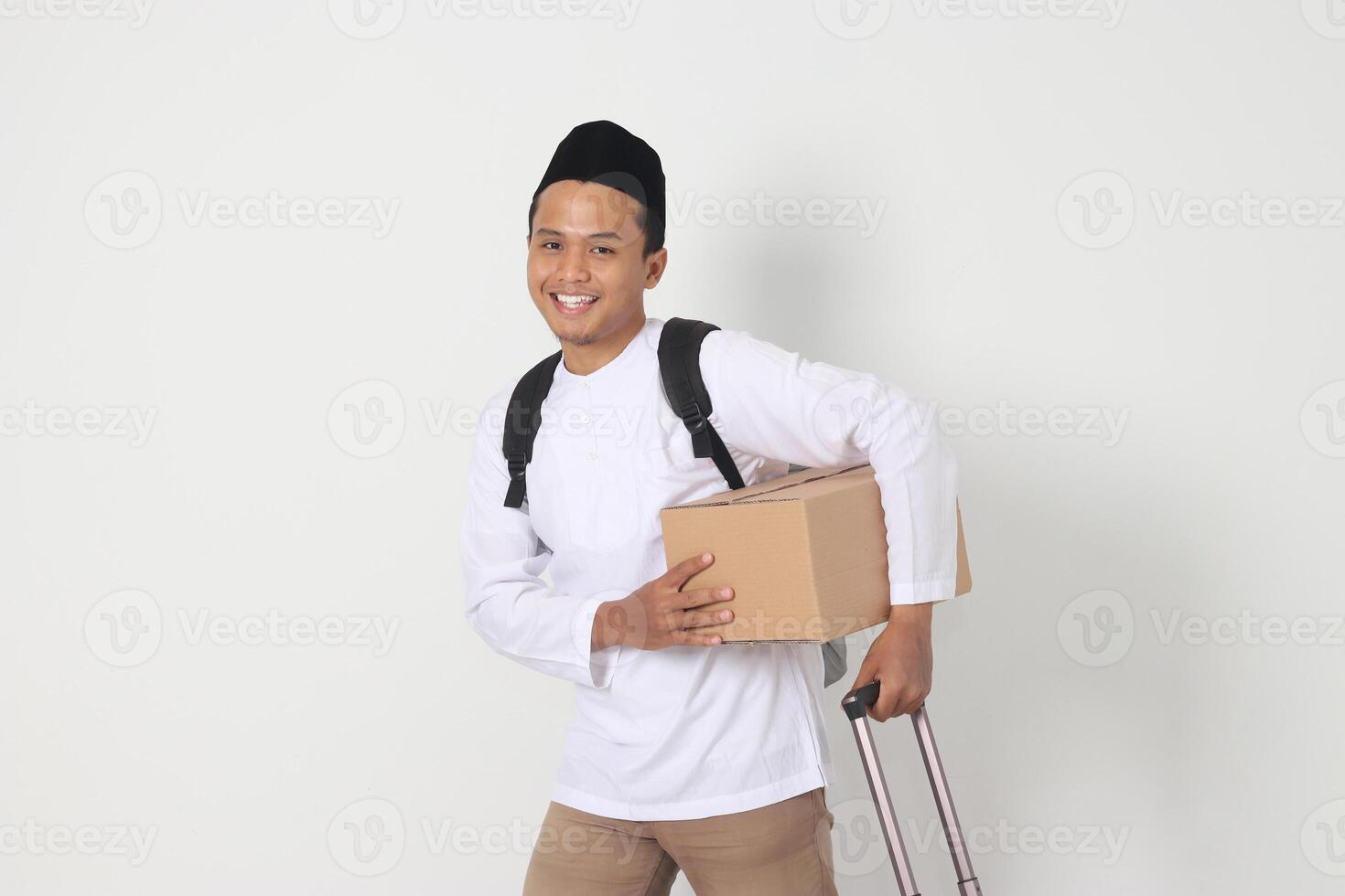 Portrait of excited Asian muslim man in koko shirt with peci carrying cardboard box and holding suitcase handle. Going home for Eid Mubarak. Isolated image on white background photo