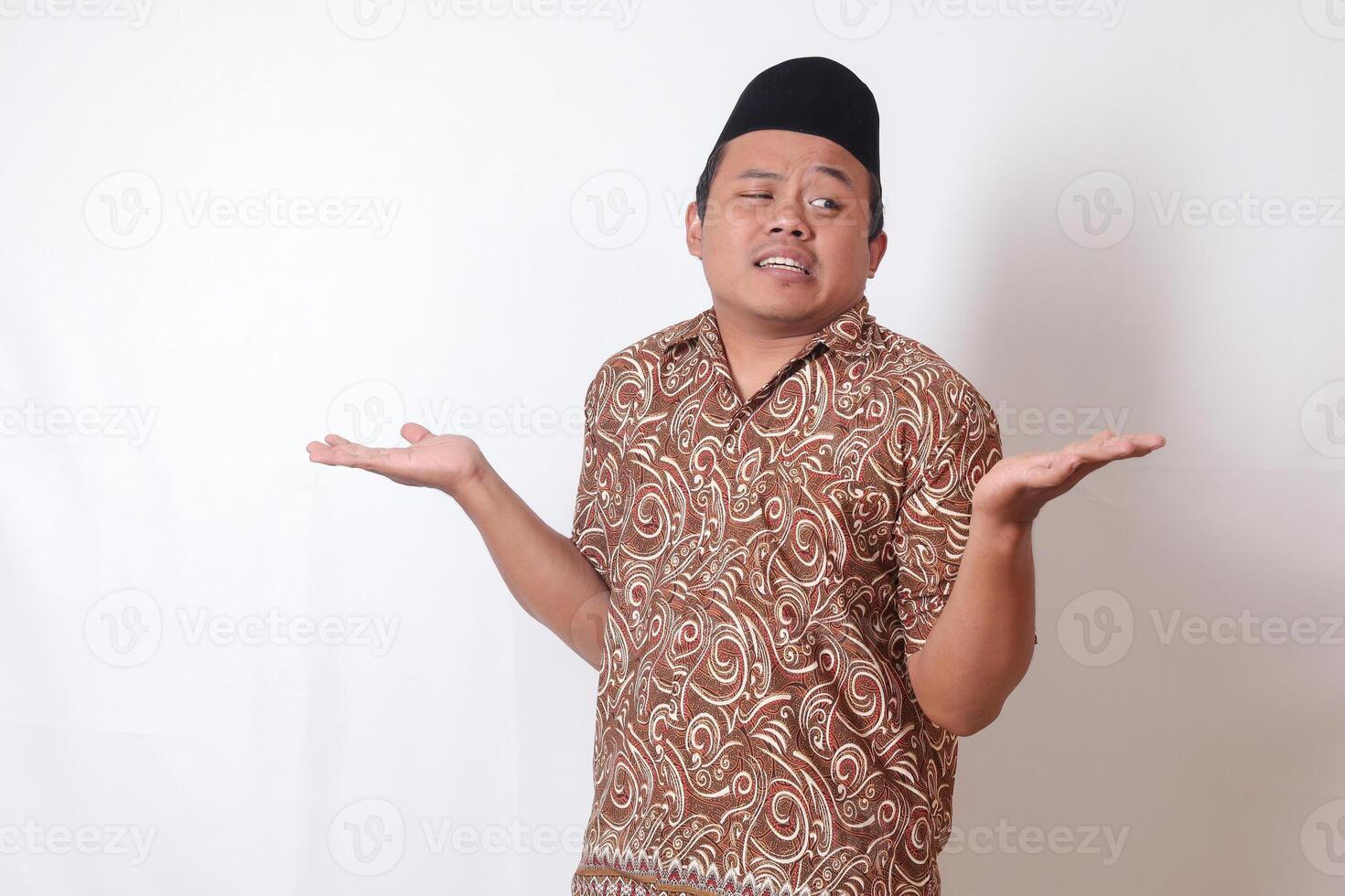 Portrait of confused Asian man wearing batik shirt and songkok with crossed hands, pointing sideways, making choice, choosing between two objects. Isolated image on gray background photo