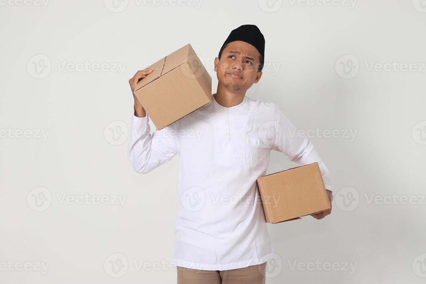 Portrait of tired Asian muslim man in koko shirt with peci having difficulty carrying cardboard. Going home for Eid Mubarak. Isolated image on white background photo