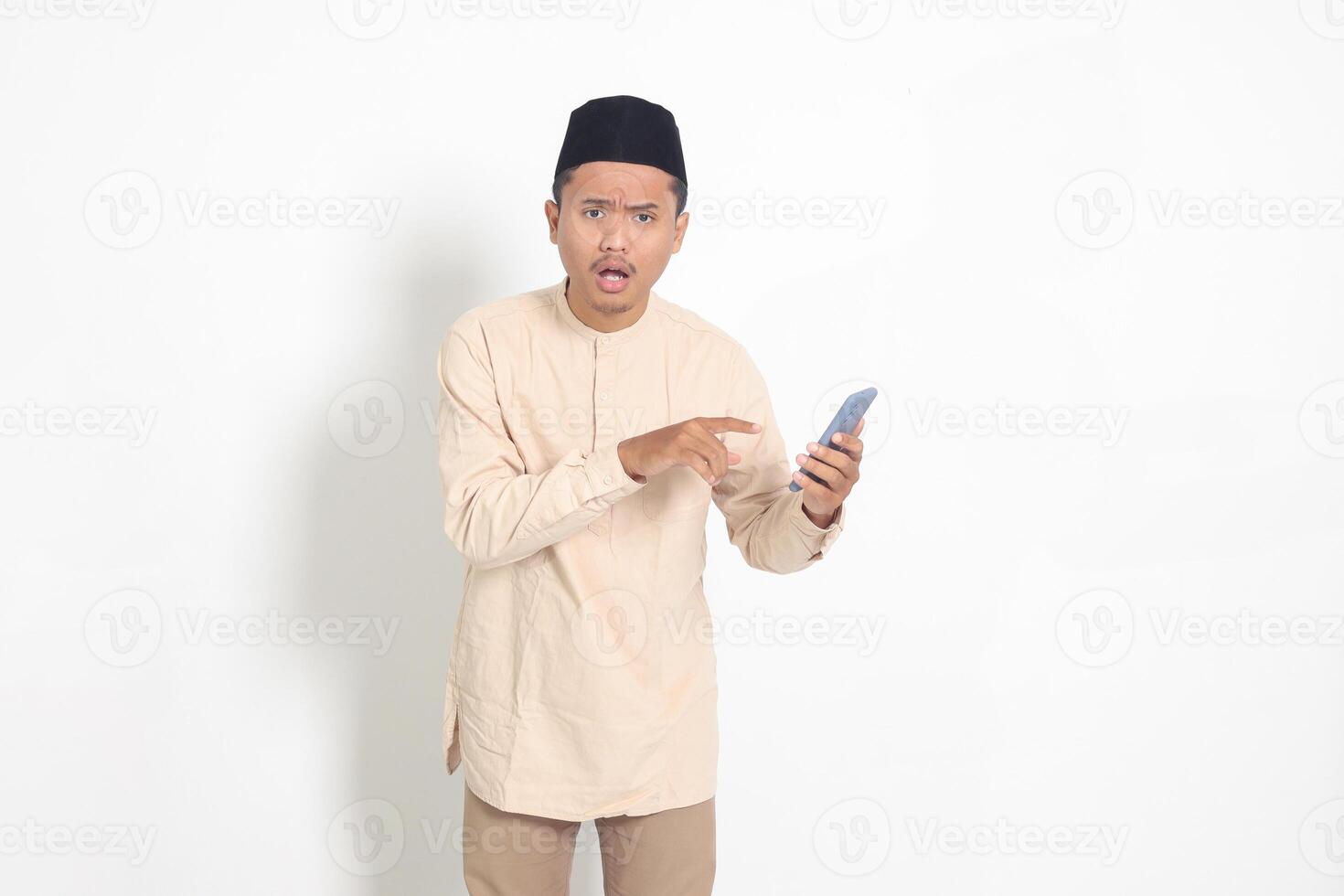 Portrait of surprised Asian muslim man in koko shirt with skullcap holding mobile phone, showing shocked face expression. Advertising and social media concept. Isolated image on white background photo