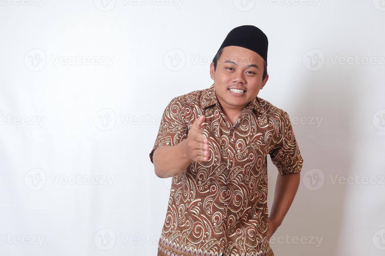 Portrait of excited Asian man wearing batik shirt and songkok reaching out his hand for a hand shake, welcoming someone. Isolated image on gray background photo