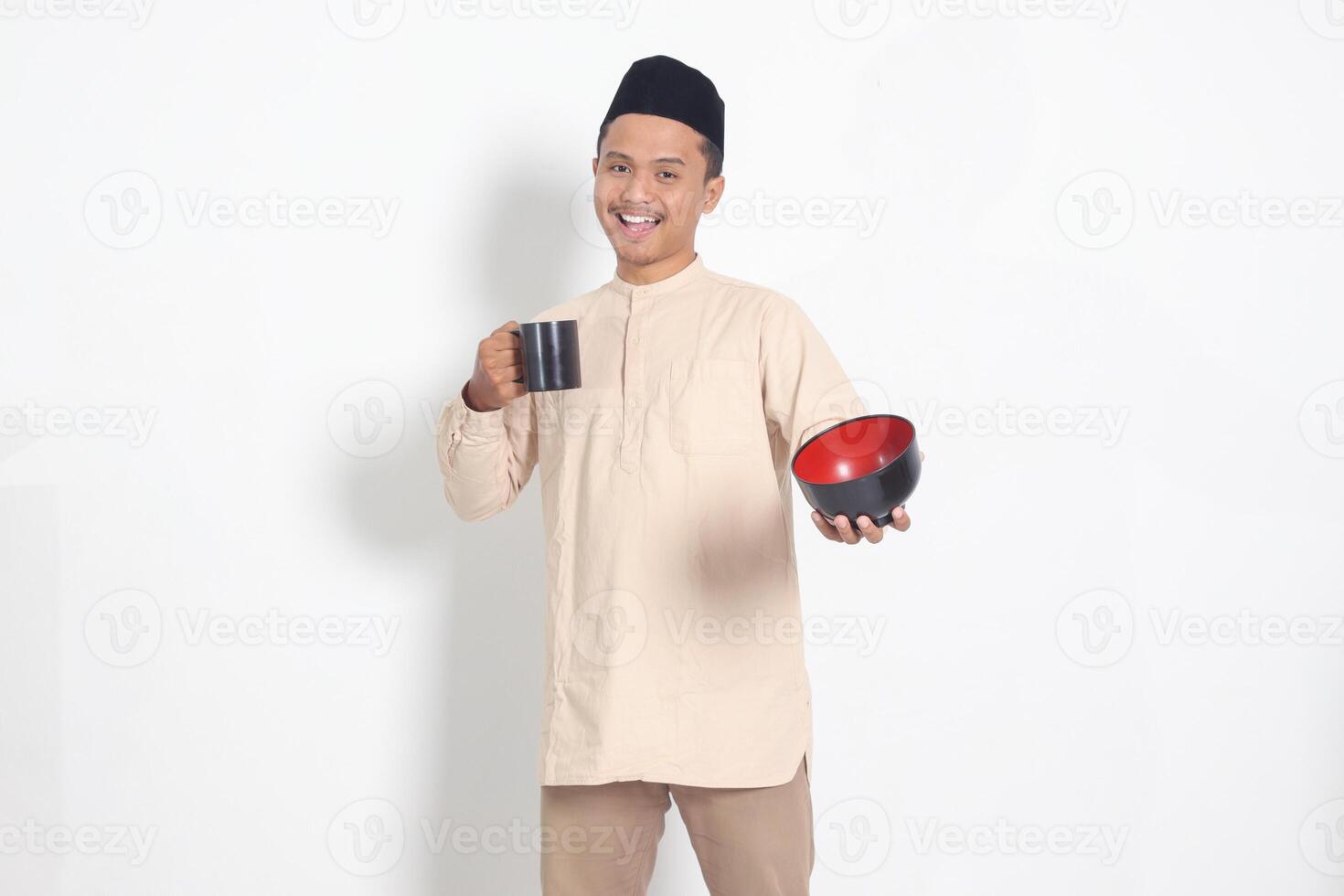 Portrait of excited Asian muslim man in koko shirt with skullcap holding a mug and empty bowl. Bowl template for food brand. Isolated image on white background photo