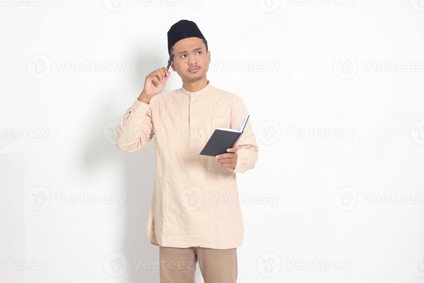 Portrait of confused Asian muslim man in koko shirt with peci reading a book, thinking about an idea with hand holding pen on chin, looking away. Isolated image on white background photo