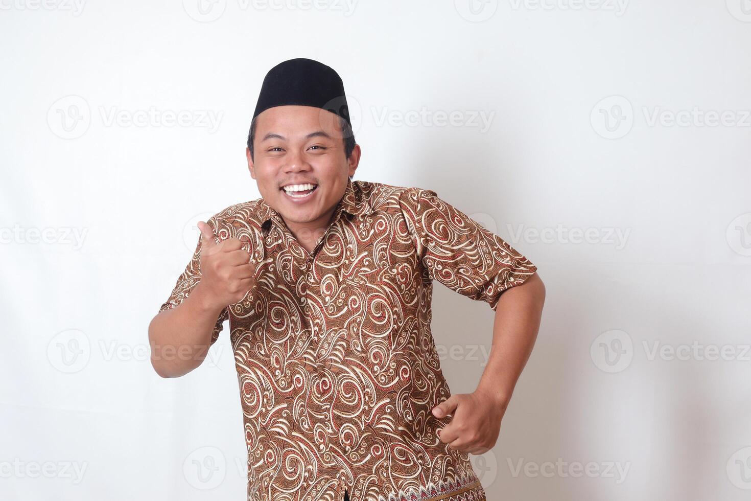 Portrait of excited Asian man wearing batik shirt and songkok smiling and looking at camera, making thumbs up hand gesture. Isolated image on gray background photo