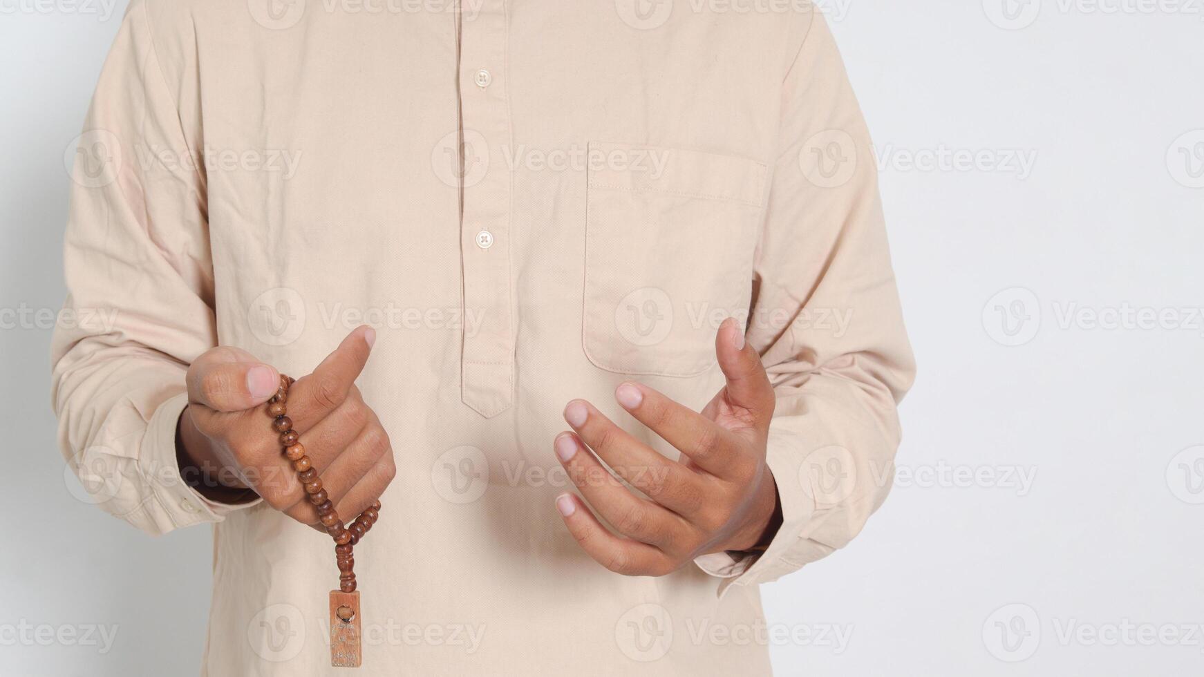 Close up portrait of religious Asian muslim man in koko shirt with skullcap praying earnestly with his hands raised, holding islamic beads. Devout faith concept. Isolated image on white background photo