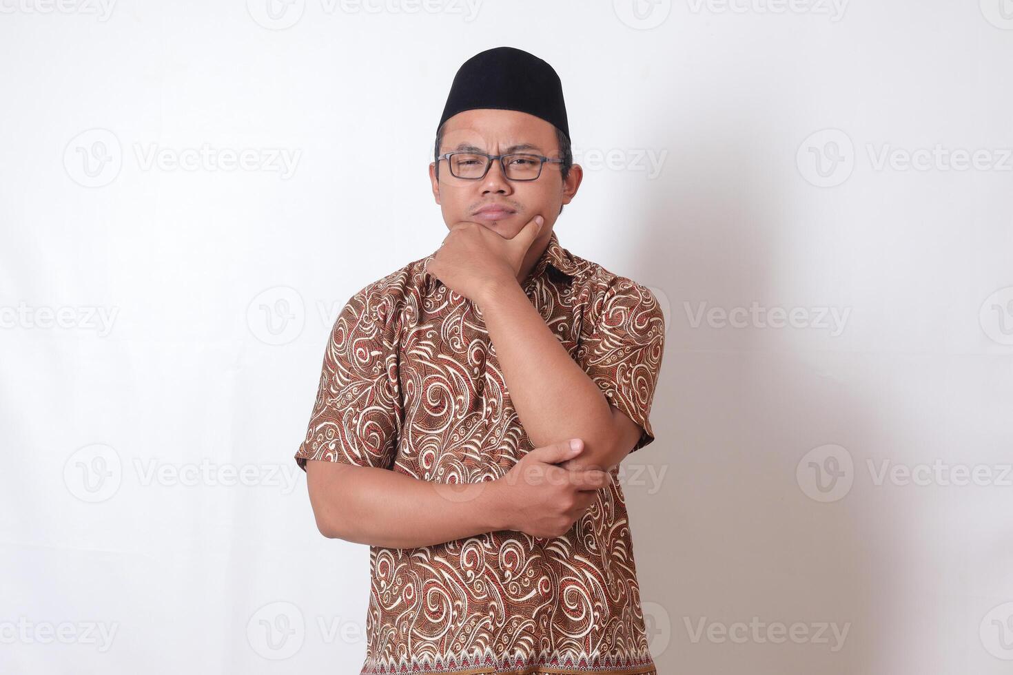 Portrait of confused Asian man wearing batik shirt and songkok standing against gray background, thinking about question with hand on chin photo
