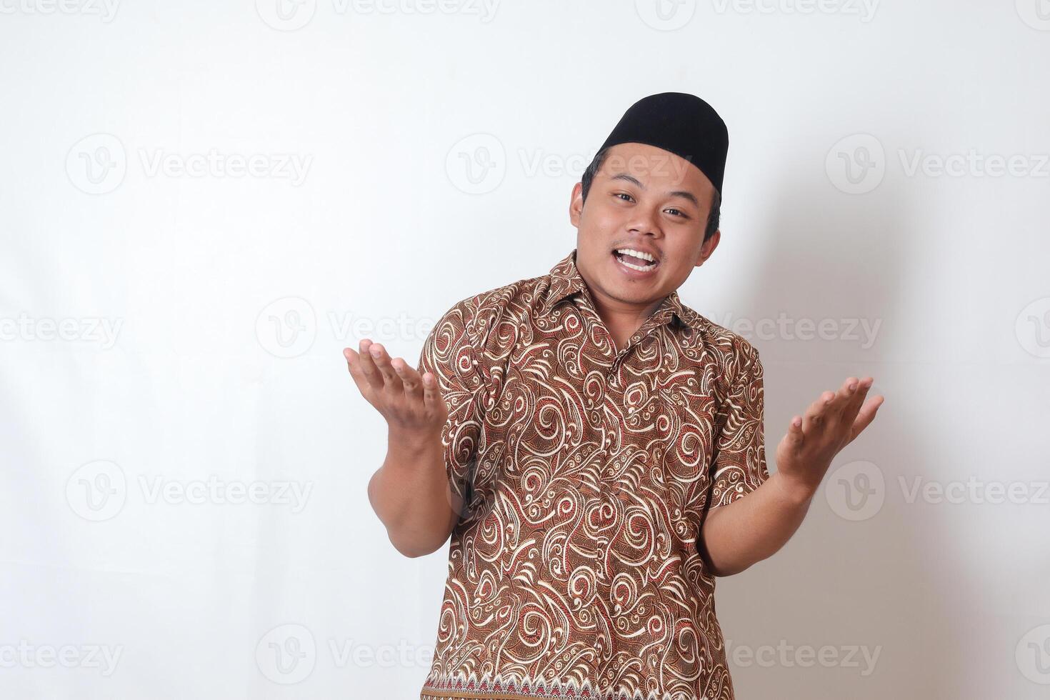 Portrait of excited Asian man wearing batik shirt and songkok spreading his hands sideways. Welcoming and greeting someone. Isolated image on gray background photo
