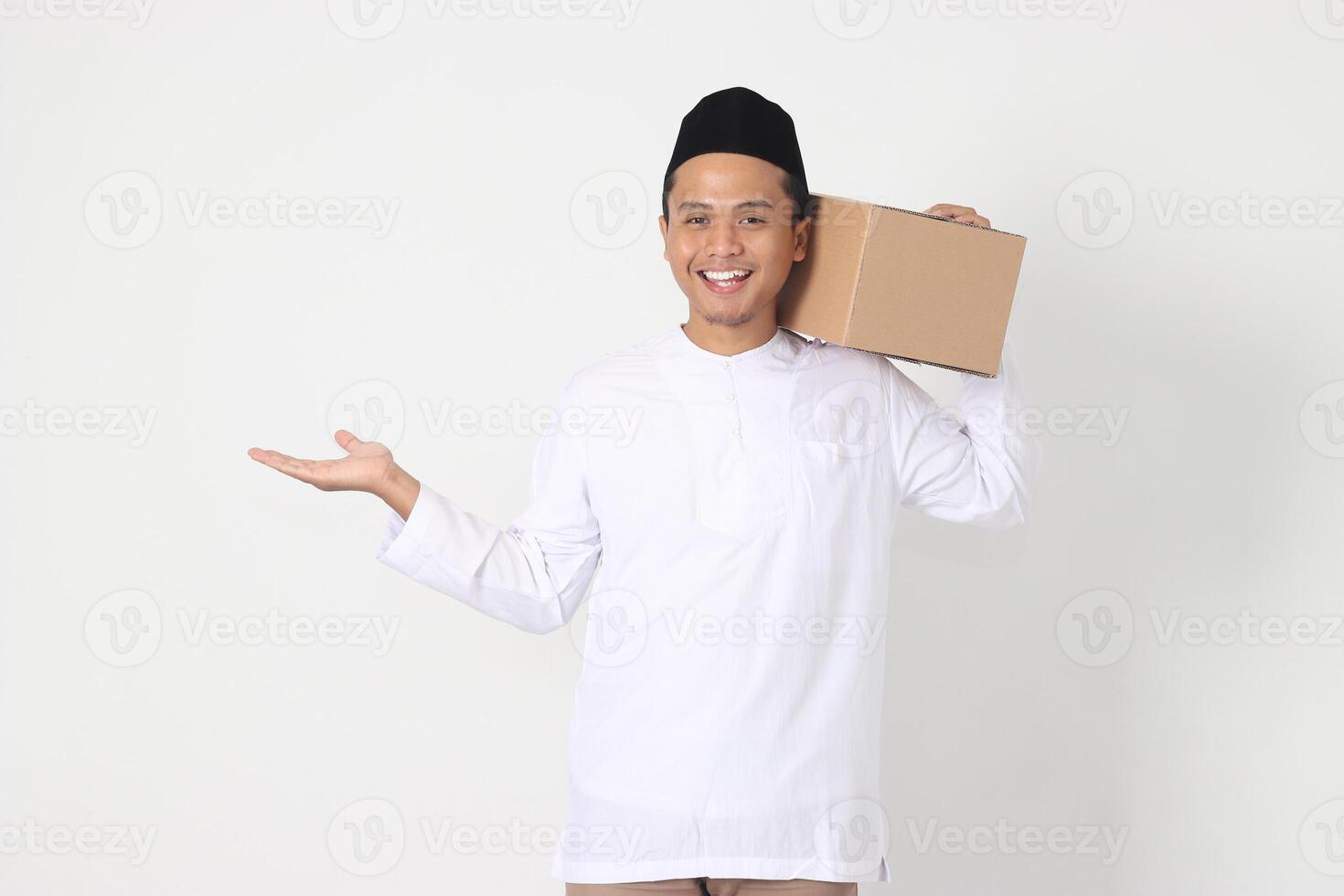 Portrait of attractive Asian muslim man in koko shirt with peci pointing to the side while carrying cardboard box. Going home for Eid Mubarak. Isolated image on white background photo