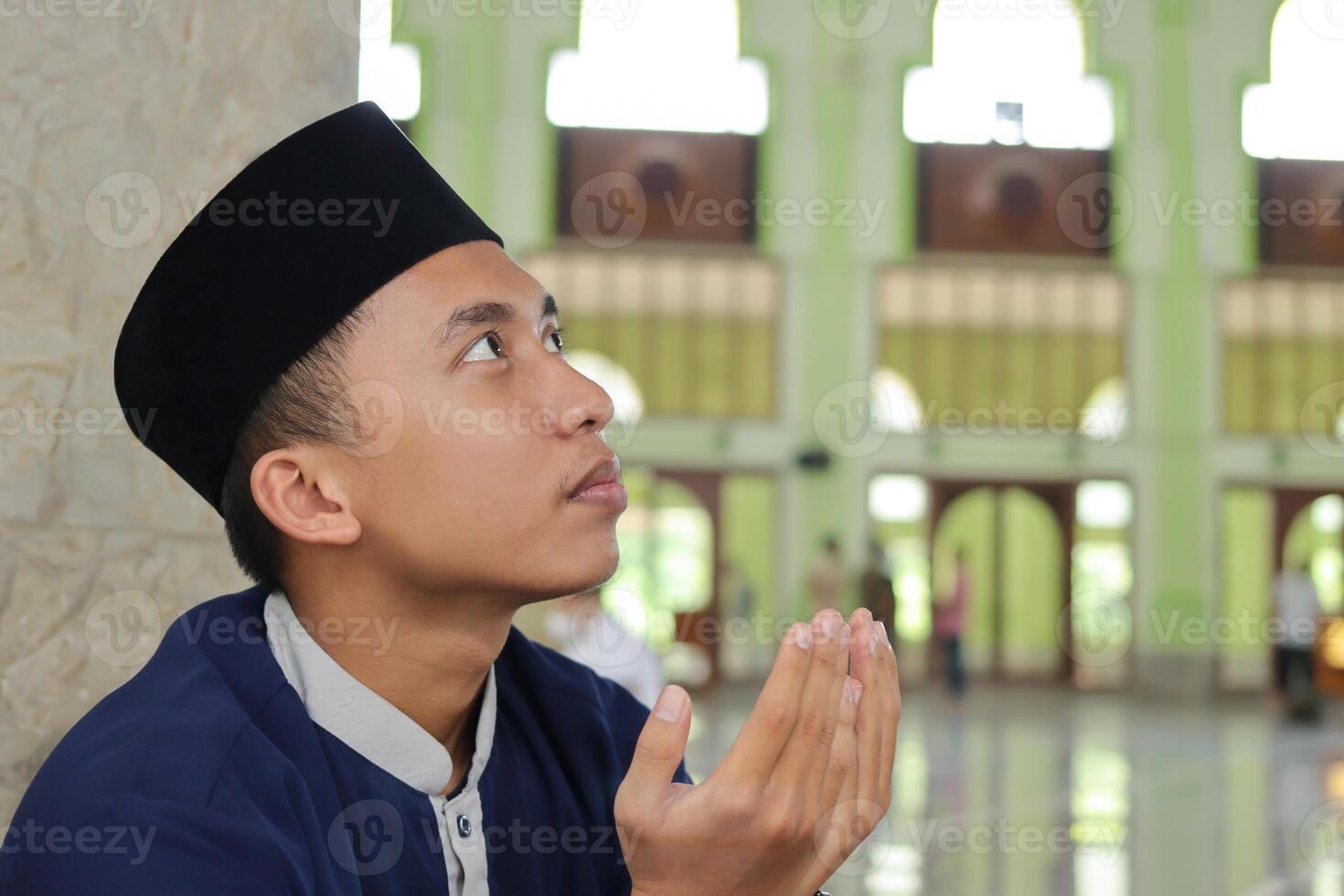 Portrait of religious Asian man in muslim shirt and black cap praying with hand raised inside the public mosque photo