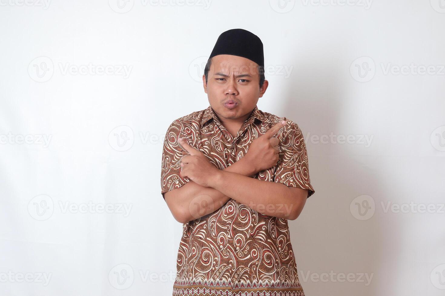 retrato de confuso asiático hombre vistiendo batik camisa y Songkok con cruzado manos, señalando oblicuo, haciendo elección, elegir Entre dos objetos. aislado imagen en gris antecedentes foto
