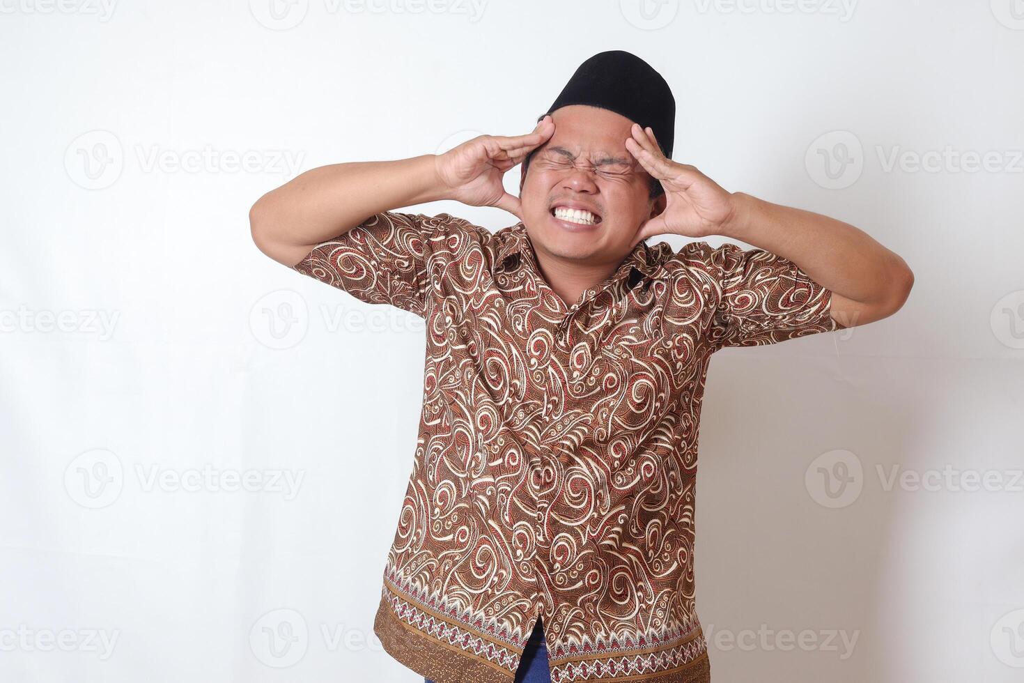 Portrait of suffering Asian man wearing batik shirt and songkok having headache while touching his forehead area. Isolated image on gray background photo